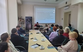 Uczestnicy spotkania siedzą przy stolikach i patrzą w kierunku siedzącej przy stoliku prelegentki. W tle kobieta wykonująca zdjęcia. 