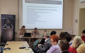 Uczestnicy spotkania siedzący przy stolikach. Dwie prowadzące kobiety siedzą przed laptopem. W tle ekran z prezentacją. 