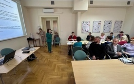 Sala konferencyjna, przy stolikach siedzą mężczyźni i kobiety. Uczestnicy spotkania patrzą w kierunku stojącej na środku kobiety z mikrofonem w ręku. 