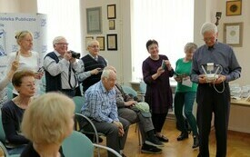 Mężczyźni i kobiety trzymający w rękach aparaty fotograficzne lub telefony patrzą w kierunku starszego mężczyzny, trzymającego w ręku urządzenie do sterowania dronem.