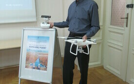 Starszy Pan w koszuli z dronem w ręku stoi obok potykacza, na kt&oacute;rym znajduje się plakat przedstawiający widok rzeki z lotu ptaka.