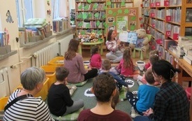 Dzieci wraz z rodzicami siedzą na dywanie na spotkaniu w bibliotece