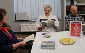 Dwie kobiety i mężczyzna siedzą przy stole, trzymają książki w rękach.