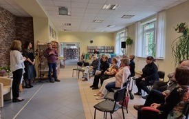 Kilkanaście osób siedzi w kole na krzesłach i słucha prowadzącej spotkanie.