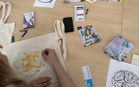 Kobieta rysuje na torbie ekologicznej kota. Wok&oacute;ł na stole szablony, kredki i pisaki do malowania na putnie. 