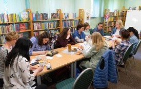 Uczestnicy warsztat&oacute;w siedzą przy wsp&oacute;lnym stole, robią notatki, rozmawiają. Wok&oacute;ł regały z książkami.