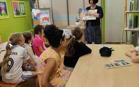 Kobieta z czarnymi włosami stoi i pokazuje palcem obrazek w otwartej książce. Przed nią przy stolikach siedzi grupa dzieci. 