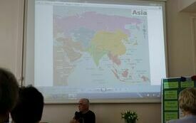 Na ekranie projekcyjnym wyświetla się mapa Azji. Poniżej starszy mężczyzna w okularach z mikrofonem.
