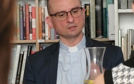 Ksiądz w koloratce, krótkich włosach i okularach. W tle regał z książkami.