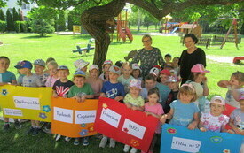 Pod drzewem grupa dzieci i dwie kobiety. Dzieci trzymają kolorowe kartony z napisem Og&oacute;lnopolski Tydzień Czytania Dzieciom 2023 z Filią nr 2