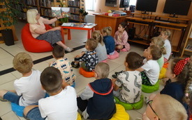 Grupa dzieci siedzących na kolorowych poduszkach w bibliotece. Dzieci wpatrzone w kobietę trzymającą w ręku otwartą książkę dla dzieci. 