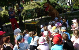 Grupa dzieci siedzących na kocyku na placu zabaw zwr&oacute;cona w kierunku kobiety z książką w ręku. Obok postać dinozaura. 