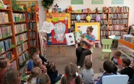 Kobieta w kolorowej bluzce pokazuje strony książki. Przed kobietą na poduszkach siedzi grupa dzieci. W tle regały z książkami. 