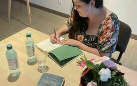 Autorka, Sylwia Winnik podpisuje książkę. Na stole dwie butelki wody, kwiaty i książka. 