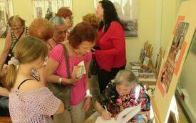 Gość spotkania starsza kobieta podczas wpisywania dedykacji do książki. Obok grupa uczestnik&oacute;w spotkania.