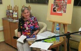 Starsza kobieta w kolorowej bluzce siedzi przy stoliku na kt&oacute;rym leżą stos książek, wydruki, woda. Obok plakat zapowiadający spotkanie. 