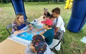 Przy stoliku z rozłożoną mapą siedzi kobieta i grupa dzieci. Kobieta wskazuje coś na mapie. 
