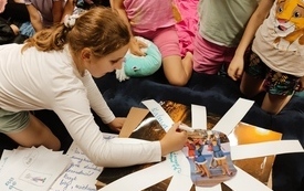 Grupa dzieci siedzi na materacu. Dziewczynka zapisuje na kartce pomysły: Jak pokonać strach?