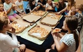Grupa dzieci je pizze pomiędzy regałami bibliotecznymi.