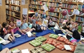 Dzieci siedzące na materacach pomiędzy regałami bibliotecznymi. Wok&oacute;ł rozłożone książki i poduszki. Dzieci trzymają w rękach Focus.