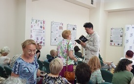 Gość spotkania stoi między publicznością z książką w ręku. Obok starsza kobieta.