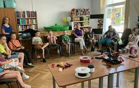 Dzieci uczestniczące w spotkaniu zgromadzone wok&oacute;ł stolika, na kt&oacute;rym stoi urządzenie do druku 3D.