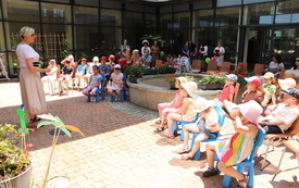 Grupa dzieci siedzi na kolorowych krzesełkach na patio przy fontannie