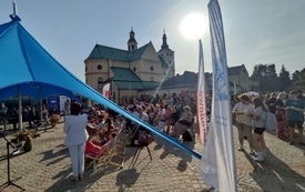 Grupa starszych i młodszych osób siedzi na krzesłach i leżakach na wybrukowanym placu, w tle za nimi budynek kościoła