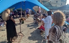 Kobieta w średnim wieku przemawia do mikrofonu przed grupą osób siedzących na krzesłach i leżakach