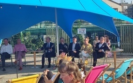 Grupa starszych i młodszych osób siedzi na krzesłach i leżakach pod dużym niebieskim namiotem