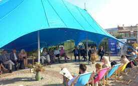 Grupa dorosłych os&oacute;b siedzi na krzesłach i leżakach pod dużym niebieskim namiotem