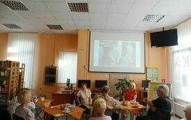Grupa dorosłych osób siedzących przy stolikach i oglądających prezentację na ekranie