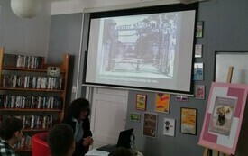 Wnętrze biblioteki. Prezentacja wyświetlana na ścianie. Kobieta prowadząca wykład i słuchacze.