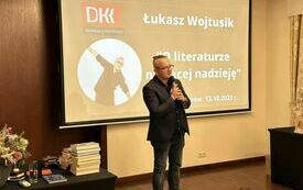 Dziennikarz Łukasz Wojtusik trzyma mikrofon i m&oacute;wi. W tle slajd prezntacji multimedialnej z tytułem &quot;O literaturze niosącej nadzieję&quot;. 