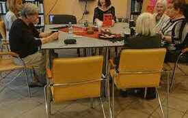 Grupa kobiet siedzi wokół stołu, w tle regały z książkami oraz ekran, na którym jest logo Dyskusyjnych Klubów Książki.