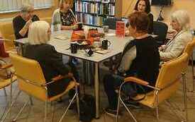 Kobiety siedzą wokół stołu, na ekranie wyświetla się logo Dyskusyjnych Klubów Książki.