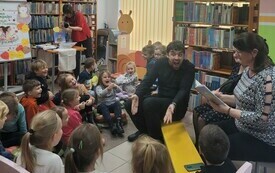 Młody mężczyzna siedzi na żółtej ławeczce, wokół grupa dzieci.