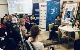 Prowadząca spotkanie kobieta w średnim wieku siedzi przy stoliku. Przed nią zgromadzona publiczność.