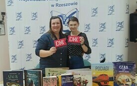 Dwie kobiety stoją obok siebie i tzrymają loga Dyskyusyjnych Klub&oacute;w Książki. Na stole wystawka z książkami. W tle logo pegaza Wojew&oacute;dzkiej i Miejskiej Biblioteki Publicznej w Rzeszowie.