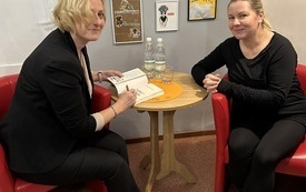 Kobiety przy stoliku. Jedna z nich podpisuje książkę. W tle kolaże w ramkach na ścianie.