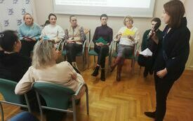 Grupa kobiet siedzi na krzesełkach. Obok stojąca kobieta.