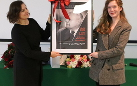 Dwie młode kobiety trzymają w rękach ramkę z plakatem z wizerunkiem Melchiora Wańkowicza.