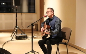 Na scenie mężczyzna w okularach, koszuli gra na gitarze i śpiewa.