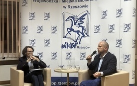 Dwaj mężczyźni siedzący w fotelach z mikrofonami w rękach. W tle baner z logotypem Wojew&oacute;dzkiej i Miejskiej Biblioteki Publicznej w Rzeszowie - pegazem.