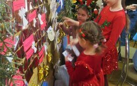 Trzy dziewczynki w świątecznych strojach stoi przy tablicy, na kt&oacute;rej zawieszono aniołki i i kolorowe karteczki