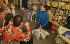 Grupa dzieci zwrócona w kierunku kobiety siedzącej na dywanie. Przy kobiecie koszyk wiklinowy. W tle regały z książkami.