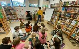 Grupa dzieci w bibliotece przygląda się kobiecie w żółtym swetrze.