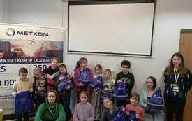 Grupa dzieci oraz trzy kobiety pozują do wspólnego zdjęcia. Dzieci trzymają w rękach granatowe worki.