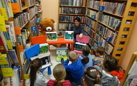Grupa dzieci siedzi pośród regałów w bibliotece.