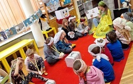 Kobieta w żółtym stroju wraz z grupą dzieci siedzi na dywanie.